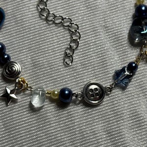 K&ouml;nnte beinhalten: Eine Perlenkette mit dunkelblauen und cremefarbenen Perlen, silberfarbenen Metallanh&auml;ngern in Form eines Sterns, einer Spirale und eines Vier-Loch-Knopfes sowie klaren Glasakzenten und einer silberfarbenen Metall-Verl&auml;ngerungskette mit Gliedern.