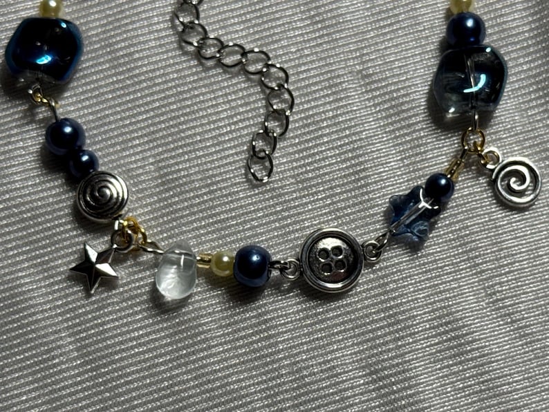 K&ouml;nnte beinhalten: Eine Perlenkette mit dunkelblauen und cremefarbenen Perlen, silberfarbenen Metallanh&auml;ngern in Form eines Sterns, einer Spirale und eines Vier-Loch-Knopfes sowie klaren Glasakzenten und einer silberfarbenen Metall-Verl&auml;ngerungskette mit Gliedern.
