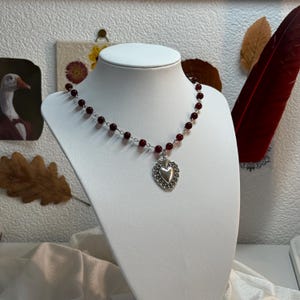 Könnte beinhalten: Eine Halskette mit einem silbernen Herz-Anhänger und einer roten Perlenkette. Der Herz-Anhänger hat ein detailliertes Design und hängt an einer silbernen Kette.