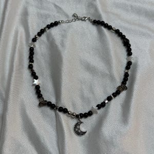 以下が含まれることがあります： 黒いビーズのネックレス、シルバーのアクセントと三日月ペンダント。ネックレスは小さな黒いビーズ、星型のシルバービーズ、シルバーチェーンが特徴です。ペンダントは詳細な三日月です。