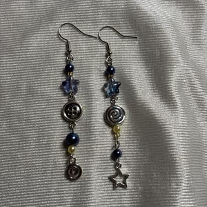 May include: A pair of dangle earrings featuring silver-toned hooks, dark blue and cream beads, translucent blue star-shaped beads, a silver-toned button charm, a silver-toned spiral charm, and a silver-toned open star charm arranged in a vertical line.