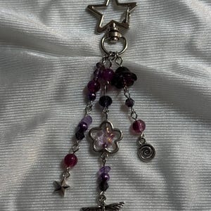 May include: A silver-toned keychain with a star-shaped top. Dangling from the keychain are purple and clear beads, a flower charm, a spiral charm, a star charm, and a dragonfly charm. The beads are various sizes and shades of purple.