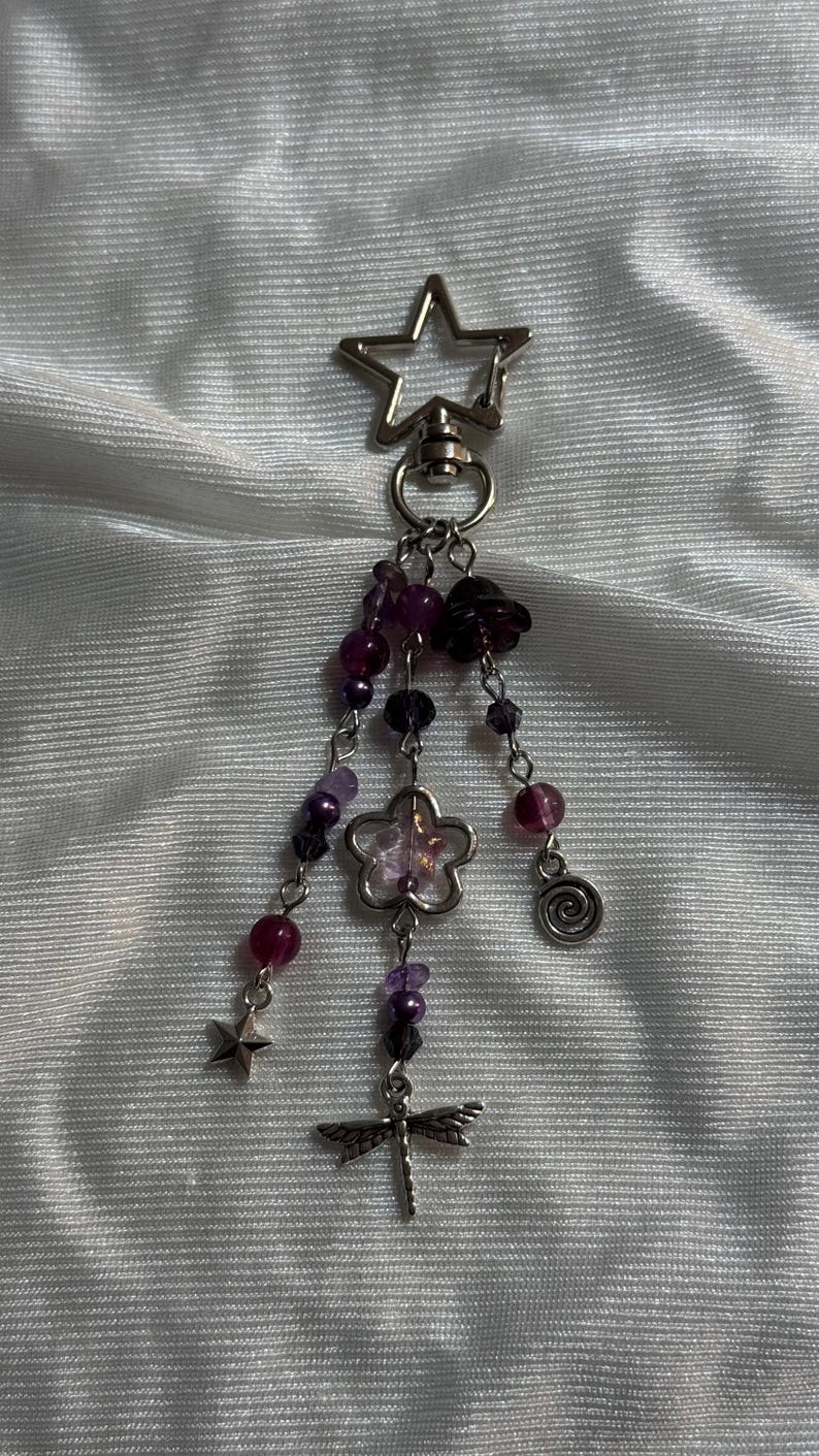 May include: A silver-toned keychain with a star-shaped top. Dangling from the keychain are purple and clear beads, a flower charm, a spiral charm, a star charm, and a dragonfly charm. The beads are various sizes and shades of purple.