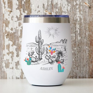 May include: White stainless steel tumbler with a black and white illustration of a desert landscape with cacti and the name "ASHLEY" printed on the bottom.