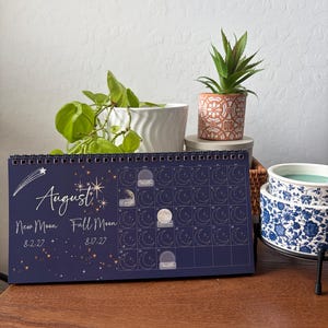 May include: A dark blue spiral-bound wall calendar for August 2027 featuring a celestial theme with stars, a shooting star, and moon phase illustrations. The text reads August, New Moon 8-2-27, and Full Moon 8-17-27, with a grid layout.