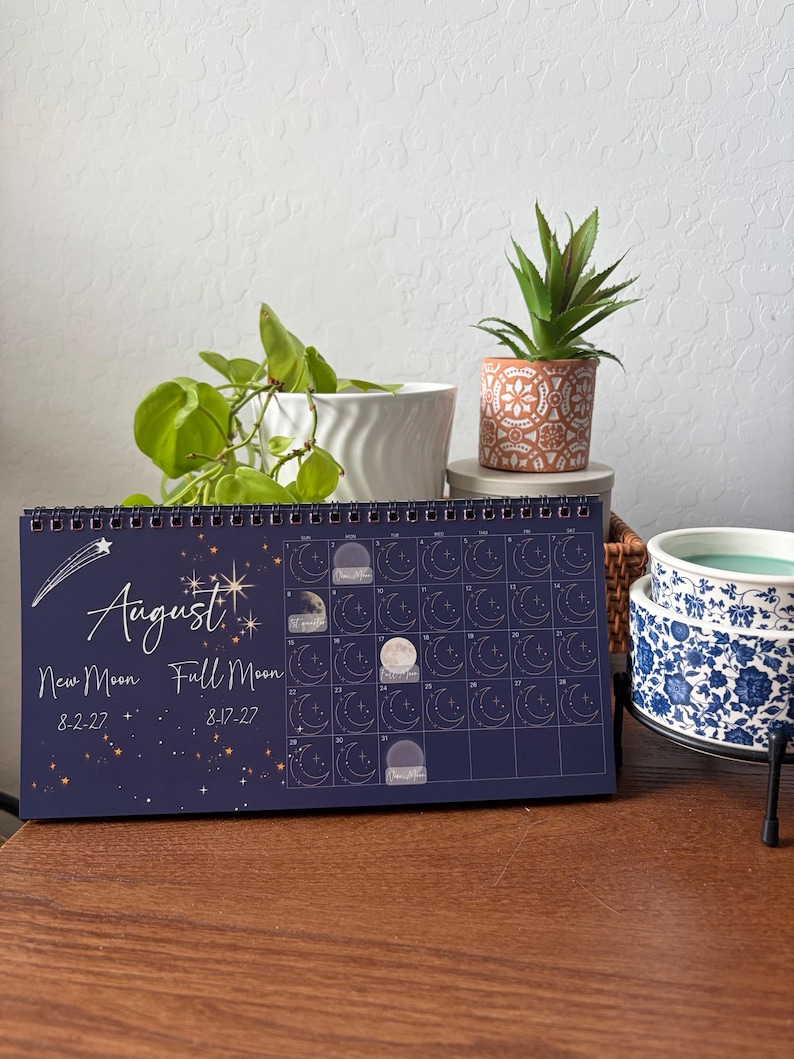 May include: A dark blue spiral-bound wall calendar for August 2027 featuring a celestial theme with stars, a shooting star, and moon phase illustrations. The text reads August, New Moon 8-2-27, and Full Moon 8-17-27, with a grid layout.
