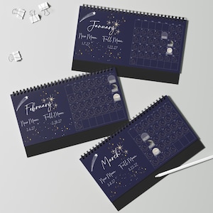 May include: Three spiral-bound desk calendars featuring a dark blue background with gold celestial illustrations of stars, shooting stars, and moon phases. Each page displays the month name, New Moon and Full Moon dates, and a grid of days.