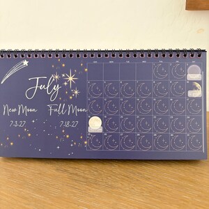 May include: A dark blue wall calendar for July featuring a white shooting star, gold stars, and a grid of thirty-one days. Each day box contains a line-art moon phase icon. Text includes July, New Moon 7-3-27, and Full Moon 7-18-27.