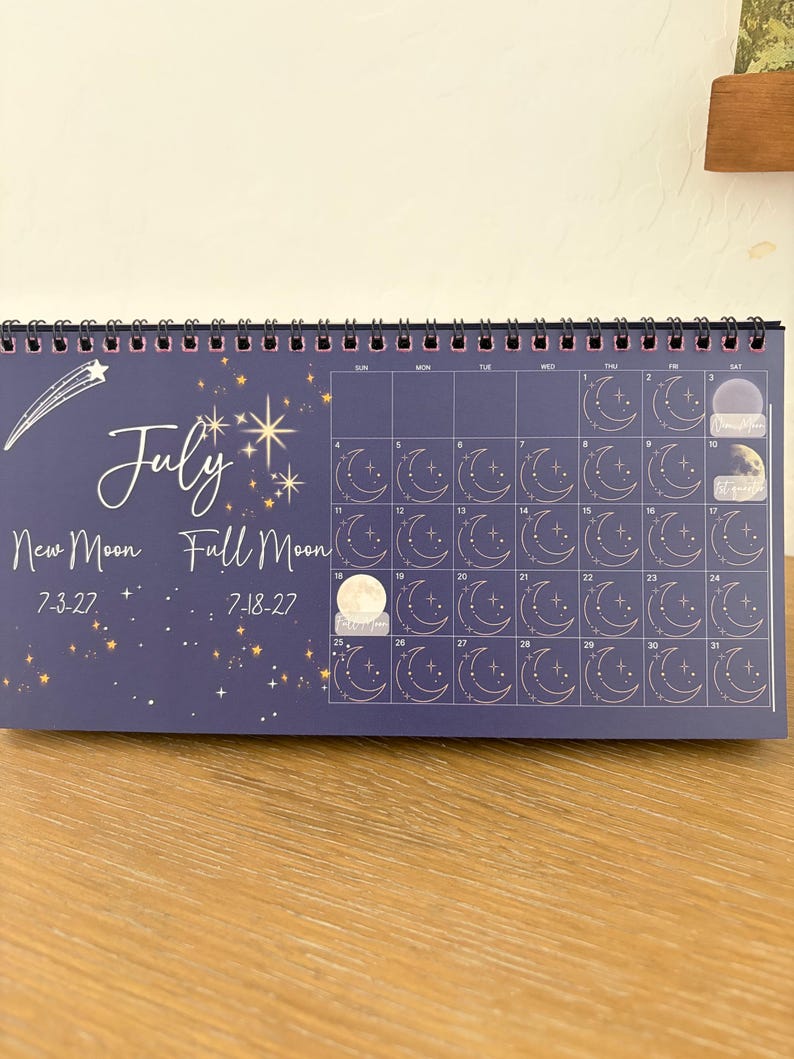 May include: A dark blue wall calendar for July featuring a white shooting star, gold stars, and a grid of thirty-one days. Each day box contains a line-art moon phase icon. Text includes July, New Moon 7-3-27, and Full Moon 7-18-27.