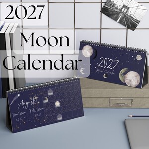 May include: A 2027 desk moon calendar with a dark blue background featuring illustrations of various moon phases, stars, and the text 2027 Moon Calendar. The calendar displays the month of August with specific dates for the new and full moon.