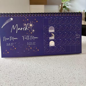 May include: A dark blue spiral-bound desk calendar for March 2027 featuring gold star illustrations, a shooting star, and moon phase icons. Text includes "March", "New Moon 3-8-27", "Full Moon 3-22-27", and "Spring equinox". Three moon stickers are applied to the grid.
