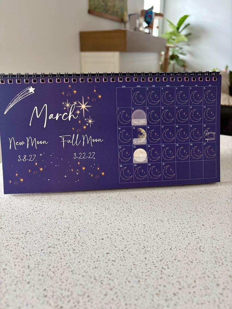 May include: A dark blue spiral-bound desk calendar for March 2027 featuring gold star illustrations, a shooting star, and moon phase icons. Text includes "March", "New Moon 3-8-27", "Full Moon 3-22-27", and "Spring equinox". Three moon stickers are applied to the grid.