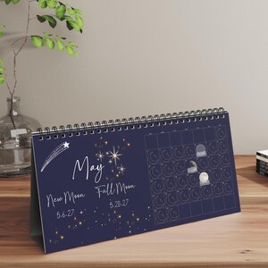 May include: A dark blue horizontal desk calendar for the month of May featuring a white shooting star, gold star illustrations, and text reading May, New Moon 5-6-27, and Full Moon 5-20-27. The calendar includes a grid with moon phase icons.