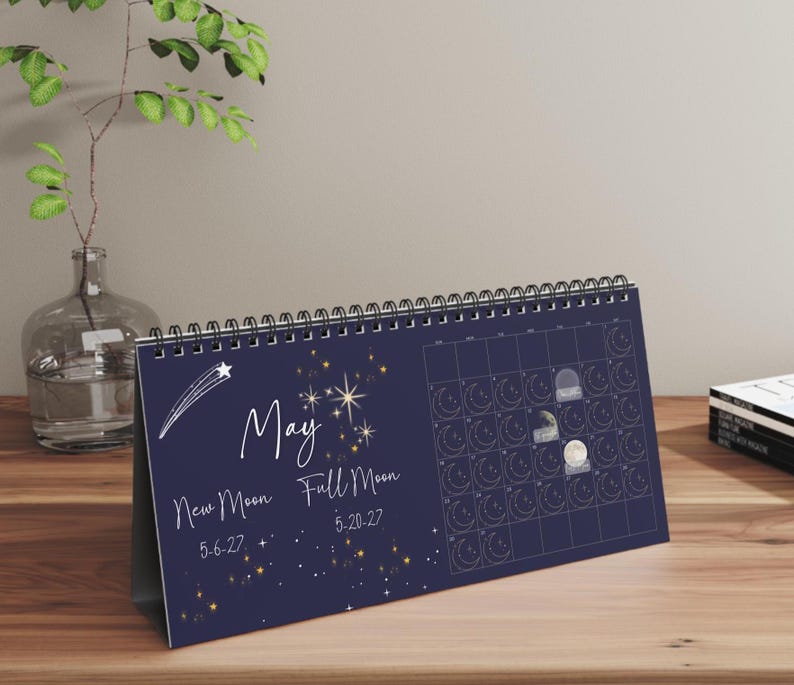 May include: A dark blue horizontal desk calendar for the month of May featuring a white shooting star, gold star illustrations, and text reading May, New Moon 5-6-27, and Full Moon 5-20-27. The calendar includes a grid with moon phase icons.