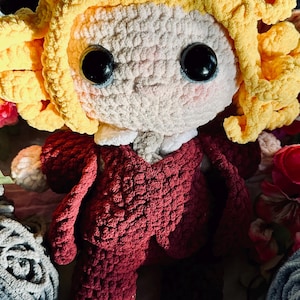 May include: A handmade crochet doll with a light complexion, large black eyes, and rosy cheeks. The doll has curly yellow hair and is dressed in a burgundy suit with a white collar. The doll is surrounded by yarn and flowers.