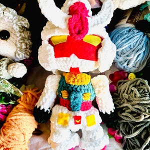 May include: A handmade, plush Gundam robot toy, primarily white with red, yellow, and blue accents. The toy is made of soft, textured yarn and features detailed stitching. Other yarn and plush toys are in the background.