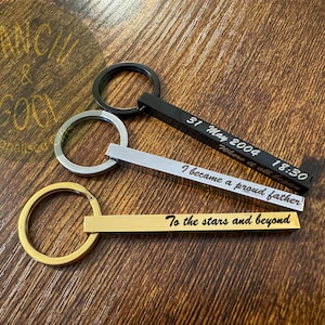 May include: Three personalised keychains with different engraved messages. The keychains are made of metal and have a silver, gold, and black finish. The messages are "31 May 2004 18:30 Where it all began", "I became a proud father", and "To the stars and beyond".