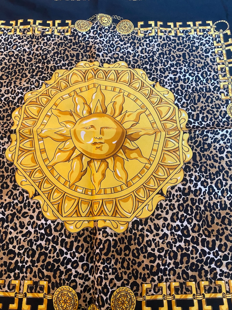 May include: A gold sun with a human face in the centre, surrounded by a gold frame and a leopard print background. The sun is set against a black background with a gold chain border.
