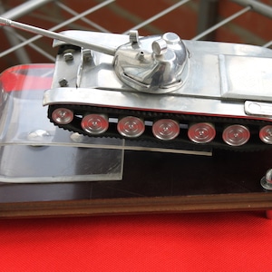 May include: A silver model of a tank on a wooden base with a clear plastic cover. The tank has a long barrel and a rounded turret. The base has four silver studs.