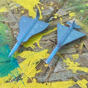 Pode incluir: Dois pequenos aviões de brinquedo azul claro com detalhes de estrelas e texto. Um é rotulado "19-144" e o outro "ЯК-25". Eles repousam sobre uma superfície de madeira salpicada com tinta amarela e verde, criando um fundo lúdico e artístico.