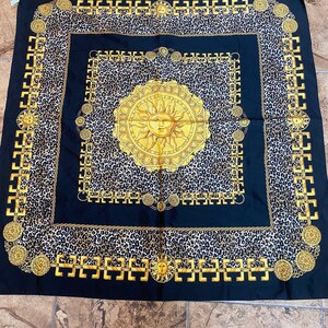 May include: A black silk scarf with a gold chain border and a leopard print background. A large gold sun with a face is in the centre of the scarf.