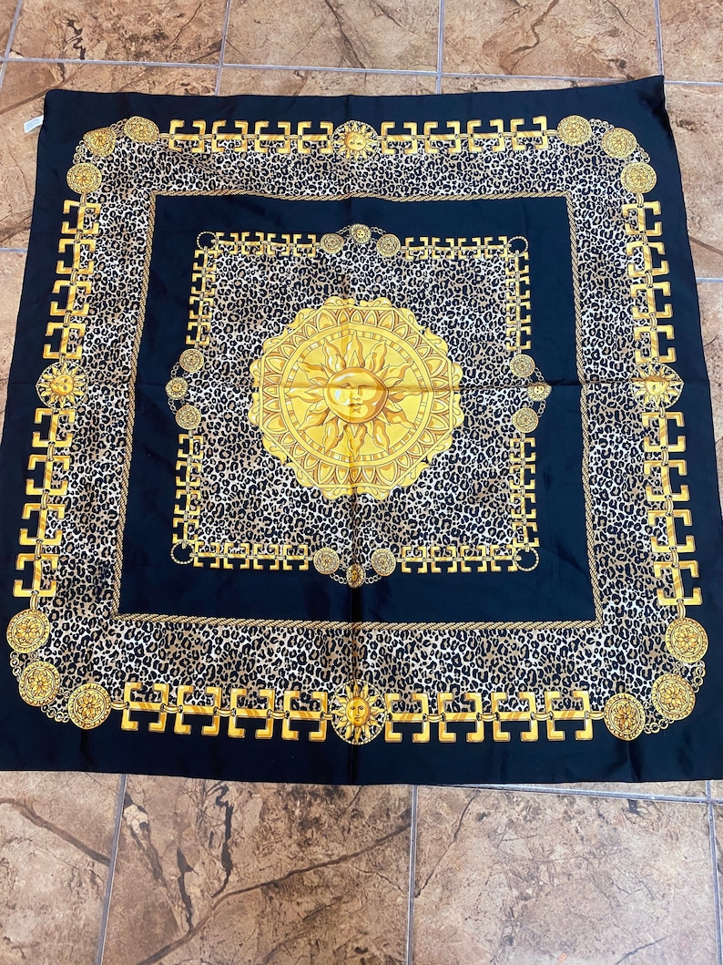 May include: A black silk scarf with a gold chain border and a leopard print background. A large gold sun with a face is in the centre of the scarf.
