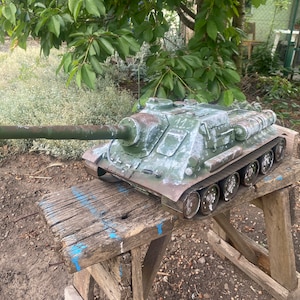 May include: A green toy tank with a long barrel, sitting on a weathered wooden stool. The tank has a camouflage pattern and appears to be made of plastic.