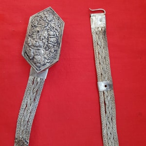 May include: A silver belt with a decorative, ornate buckle. The buckle is octagonal and features a detailed design of a dragon or other mythical creature. The belt is made of braided silver chain.
