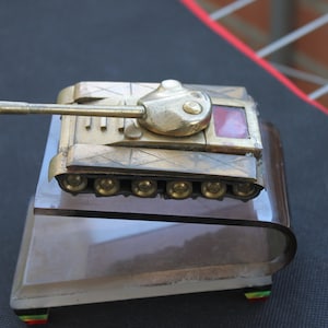 May include: A gold-coloured metal tank lighter with a red glass window on the side. The tank is sitting on a clear glass base.