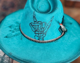 May include: A turquoise blue cowboy hat with a black skeleton hand making the 'I love you' sign on the crown. The hat has a brown band and a black feather.