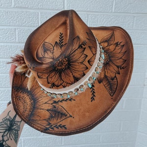 May include: Brown cowboy hat with a sunflower design. The hat features a decorative band with turquoise beads and sequins. The brim and crown are adorned with a detailed black sunflower illustration and leaf accents. A small bundle of dried flowers and feathers is attached to the side.