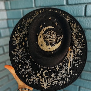 May include: Black fedora hat with gold celestial design. The hat features a crescent moon, stars, and floral accents. The brim is decorated with gold leaves and stars. A stylish accessory for any occasion.