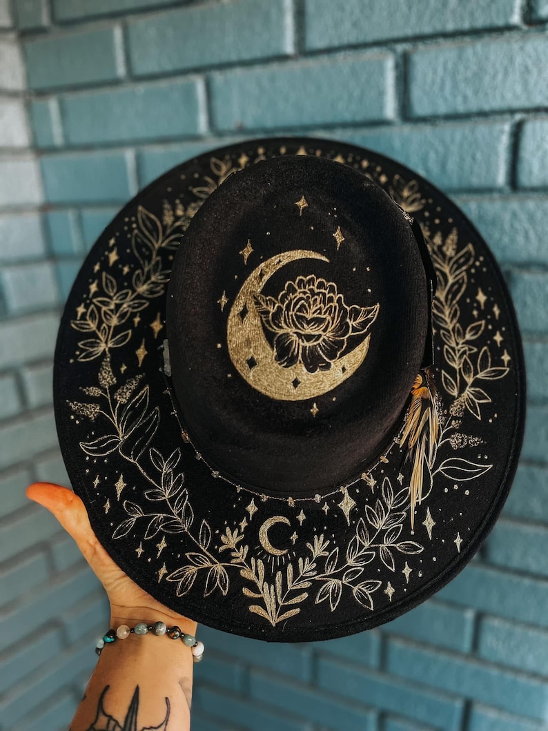K&ouml;nnte beinhalten: Schwarzer Fedora-Hut mit goldenem Himmelsdesign. Der Hut hat einen Mond, Sterne und florale Akzente. Der Rand ist mit goldenen Bl&auml;ttern und Sternen verziert. Ein stilvolles Accessoire f&uuml;r jeden Anlass.