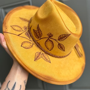 以下が含まれることがあります： レモンの枝のデザインが施された黄色のスエードフェドーラハット