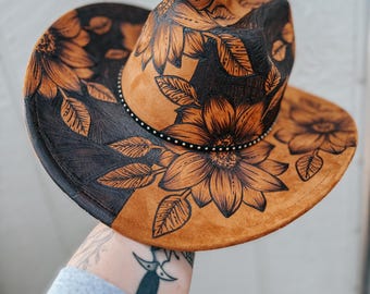 May include: A tan cowboy hat with a floral design. The hat features a black and tan color scheme with detailed sunflower and leaf illustrations. The brim is decorated with small silver studs.