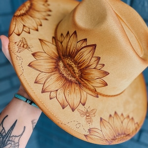 Peut inclure: Un chapeau fedora en feutre beige avec un motif de tournesol et des accents d'abeilles. Le chapeau a un large bord et une couronne haute.