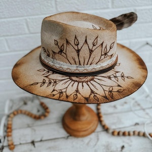 May include: A brown felt cowboy hat with a sunflower design burned into the crown. The hat has a brown leather band with a white trim and a small feather on the side.