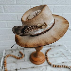 May include: A brown felt cowboy hat with a distressed finish. The hat has a brown leather band with a white feather accent. The hat is displayed on a wooden stand.