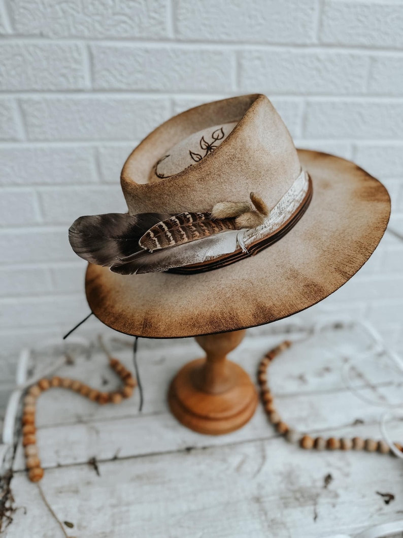 May include: A brown felt cowboy hat with a distressed finish. The hat has a brown leather band with a white feather accent. The hat is displayed on a wooden stand.