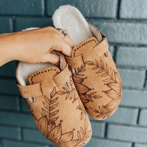 Pode incluir: Um par de tamancos de camurça marrom com um design floral. Os tamancos têm uma tira na parte superior e um forro felpudo.