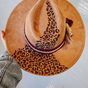 May include: A tan suede fedora hat with a leopard print design. The hat features a maroon and white band, embellished with rhinestones and a decorative feather and tassel. The brim measures approximately 7.6 cm.