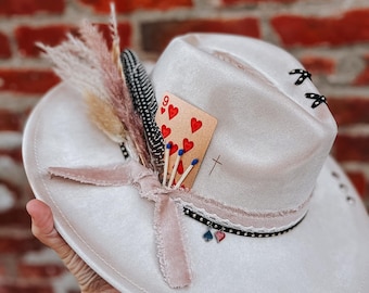 May include: A white felt cowboy hat with a pink ribbon, a feather, and a playing card with the nine of hearts. The hat has black stitching and a black beaded trim.