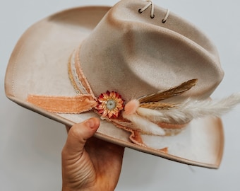 Pode incluir: Um chapéu de cowboy bege com um adorno floral decorativo e penas. O chapéu apresenta uma aba larga e uma fita com textura de veludo. O chapéu é segurado contra um fundo branco.