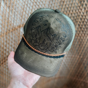 Peut inclure: Casquette de camionneur vert olive avec un motif de montagne et de fleurs. La casquette présente un graphisme noir de montagnes, de fleurs et d'un croissant de lune. Une bande marron et noire avec des points dorés orne le bord. L'arrière de la casquette est en mesh.