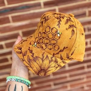 May include: A brown suede baseball cap with a hand-painted design of a honeycomb, bees, and sunflowers.