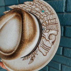 May include: A white cowboy hat with a brown skull and feather design. The hat has turquoise accents.