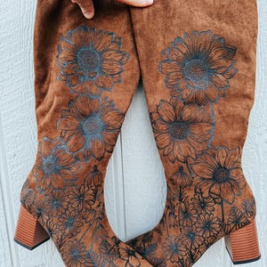 May include: Brown knee-high boots with a floral design. The boots have a block heel and are decorated with a detailed black and blue floral pattern. The boots are made of a suede-like material.