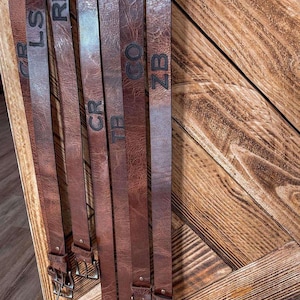 May include: A set of six brown leather belts with different initials engraved on them. The belts are hanging on a wooden surface.