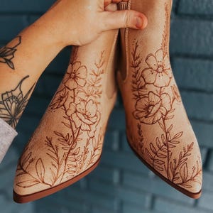 May include: Beige ankle boots with floral designs. The boots have a pointed toe and a low heel. The floral design is a darker shade of brown and covers the front and sides of the boots. The boots are held up by a person's hand.