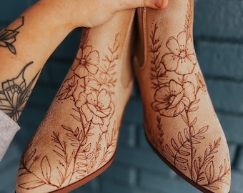 Pode incluir: Botas de tornozelo bege com desenhos florais. As botas têm bico fino e salto baixo. O desenho floral é de um castanho mais escuro e cobre a frente e os lados. As botas são seguradas por uma mão.